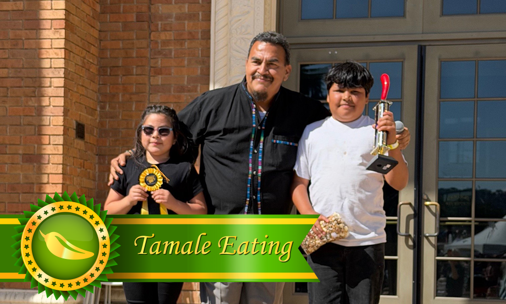 Group photo of the 2026 Tamale Festival Tamale Eating Contest winners (age 11 and under), with one person holding a trophy and another holding a ribbon, standing on either side of the event MC.